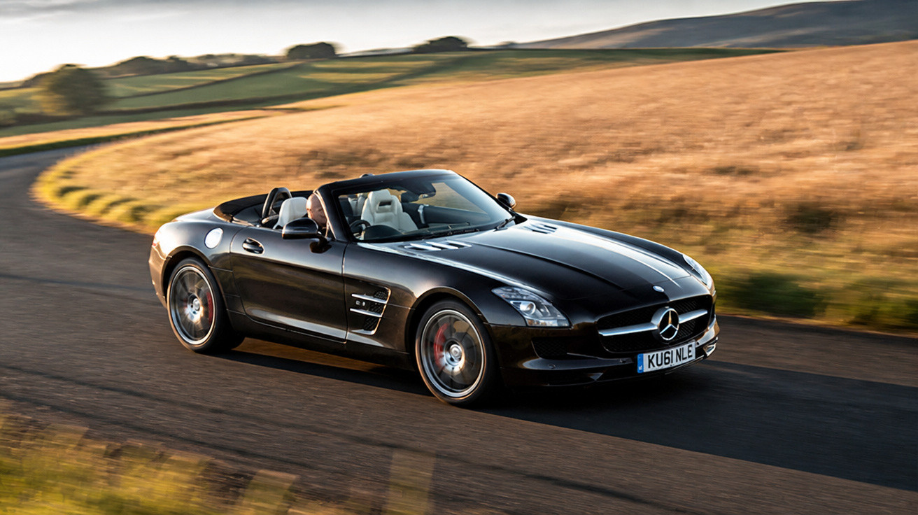 Black Mercedes convertible driving on countryside road with golden fields and hills in the background.